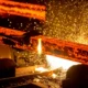 Processo de corte de barra de aço incandescente em siderúrgica, com faíscas e metal líquido.
